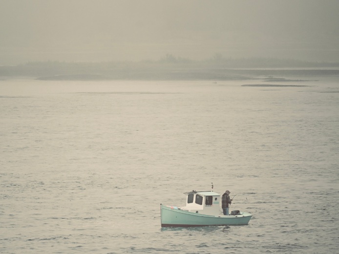 A fisherman in a small pale-green boat on a wide, misty river.
